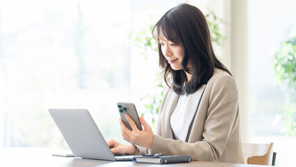 スマートフォンを持ちながらノートパソコンを見る女性ビジネスパーソン, Businesswoman holding smartphone while looking at laptop © Trickster*