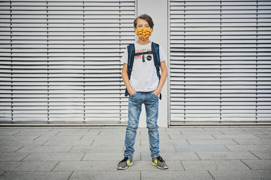 Boy with schoolbag wearing mask standing in front of building