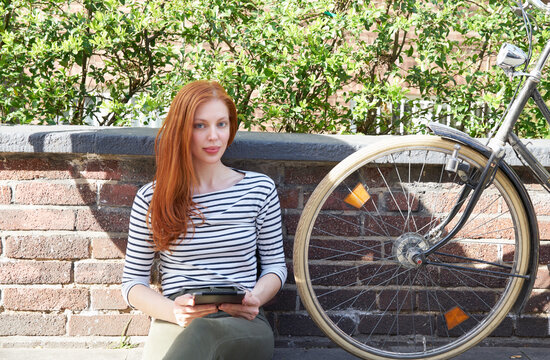 Portrait of redheaded woman with tablet and bicycle sitting on a wall