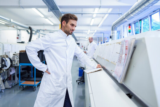 Young male technician in lab coat looking at reports on machinery