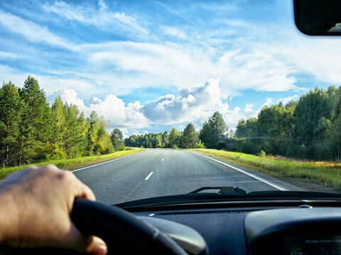 view through car windshield of natural landscape with green forest, white clouds in blue sky. Hand of woman on steering wheel of car. Female traveler