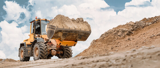 A powerful excavator loader lifts a heavy load of soil against a backdrop of fluffy clouds. Soil...