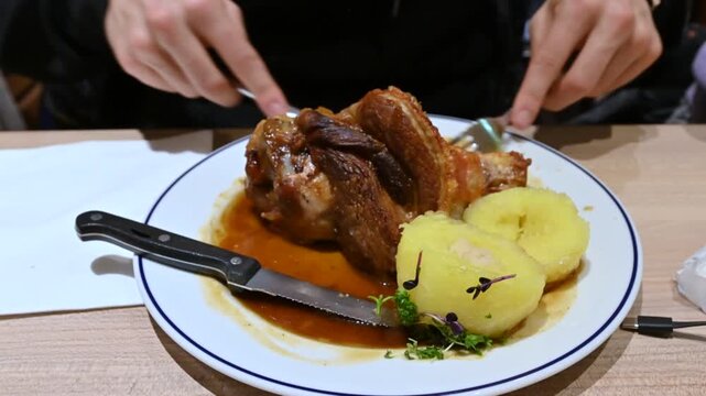 Munich, germany, december 27, 2025. Man's hands using a fork and knife to cut a large piece of roasted pork knuckle. Delicious traditional german schweinshaxe meal served on a plate