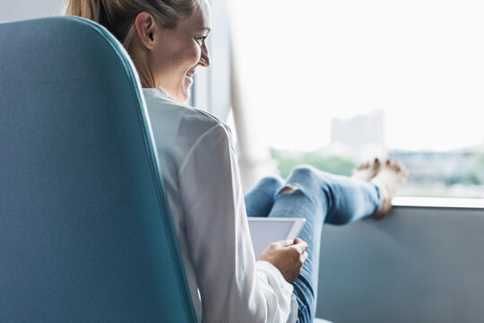 Smiling woman sitting by the window with feet up using tablet