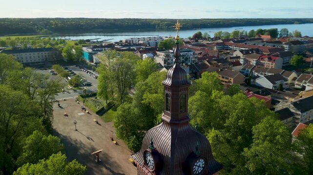 Drone circles historic V&auml;nersborg church clock tower revealing copper dome golden spire red brick facade surrounding city apartments rooftops green trees and panoramic Lake Vanern backdrop