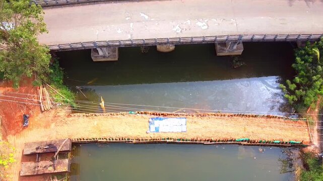This drone footage provides a high-angle, top-down perspective of a temporary earthen bridge or dam construction site alongside an existing concrete bridge. The camera slowly zooms in on the dirt path