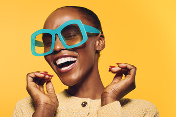 Happy African American woman with dark skin wearing large blue sunglasses and beige sweater smiles...