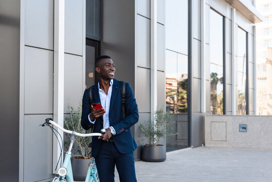 Smiling African american businessman holding smartphone while walking with bicycle outside office