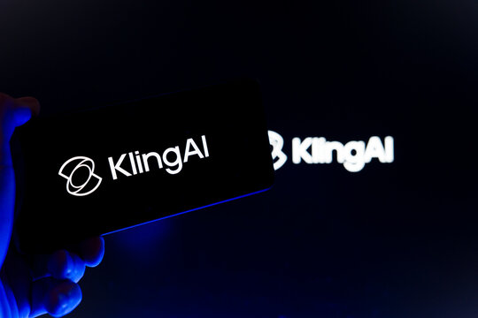 Hand holding smartphone displaying Kling AI logo on screen with blurred website interface in background, generative artificial intelligence and high-fidelity video creation technology concept