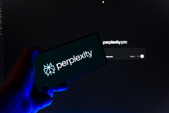 Hand holding smartphone displaying Perplexity AI logo on screen with blurred Perplexity Pro website in background, generative artificial intelligence search engine and research tool concept