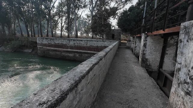 Wide-angle view of a rural irrigation canal with a small brick masonry check dam and rushing white water in a forested area of Haryana, India.