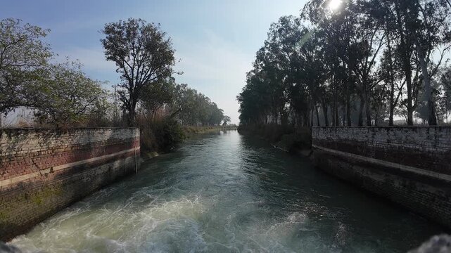 Wide-angle view of a rural irrigation canal with a small brick masonry check dam and rushing white water in a forested area of Haryana, India.
