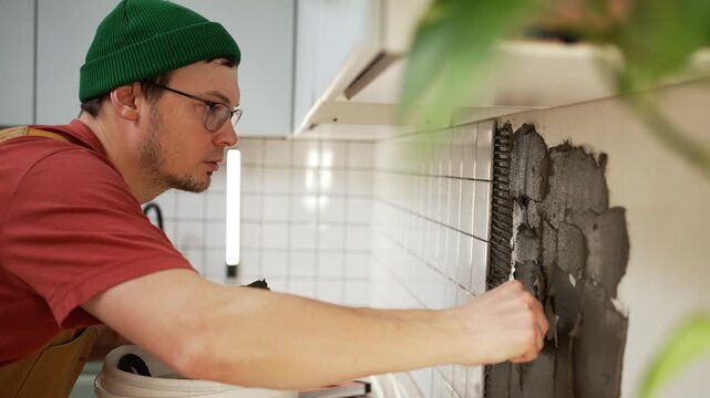 Concentrated worker meticulously installing small white ceramic tiles on a kitchen wall, applying dark gray adhesive with a special notched trowel during a diy home renovation