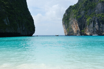 Obraz premium Tropical beach scene with turquoise water, limestone cliffs, and boats anchored near the shore under a partly cloudy sky in a serene coastal environment