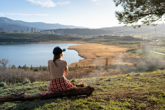 Place of power and relaxation, young woman nature explorer resting on log near fireplace in mountains, enjoying spring lake surrounded by hills and valleys, outdoor adventures on weekend. Time alone.