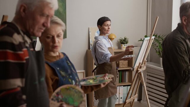 Group of Caucasian senior students in aprons painting on canvas and sharing ideas in modern art studio