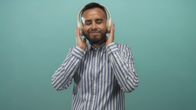Man hispanic young in striped shirt holding beige over ear headphones to ears in teal studio while hands cup headphones and eyes closed; music relaxation.