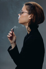Obraz premium Young woman vaping electronic cigarette with gray background in dark outfit, contemplative mood, modern lifestyle concept.