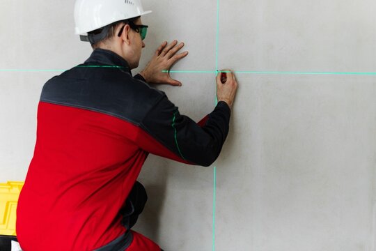 Technician Marking Wall At Intersection Of Green Laser Lines. Layout Work For Tiling Or Fixtures Installation.