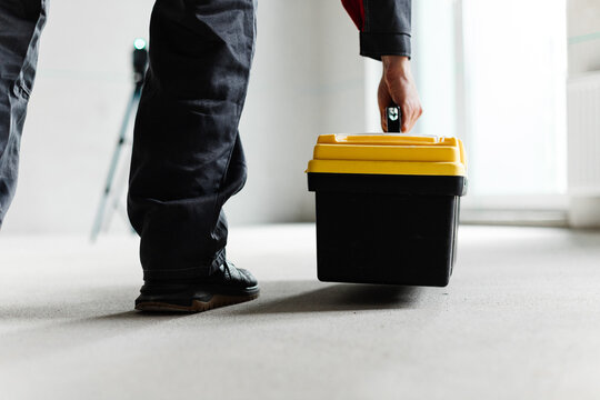 Low Angle View Of Worker Lifting Tool Case With Laser Level In Background. Construction Workflow In Raw Space.