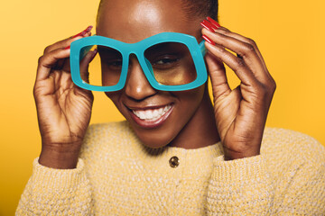Happy dark skinned African woman in yellow sweater holds oversized blue glasses on yellow...