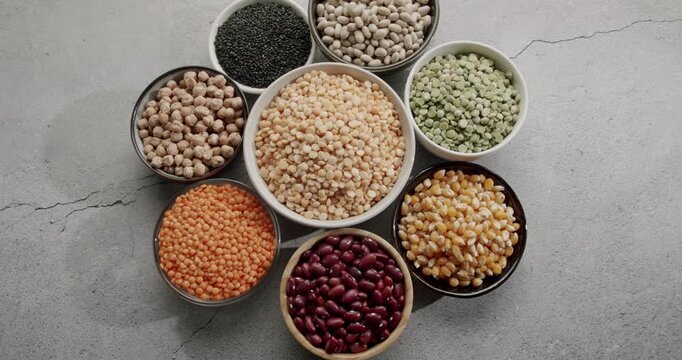 Top-down slow motion view of various dried legumes, beans, and grains rotating on a textured grey background, showcasing a rich variety of plant-based protein sources for healthy eating.