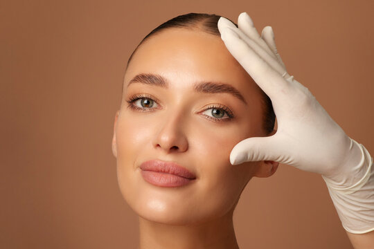 Modern aesthetic medicine. Close up of hand in glove touching young woman's face, dermatologist examining skin, standing over beige background