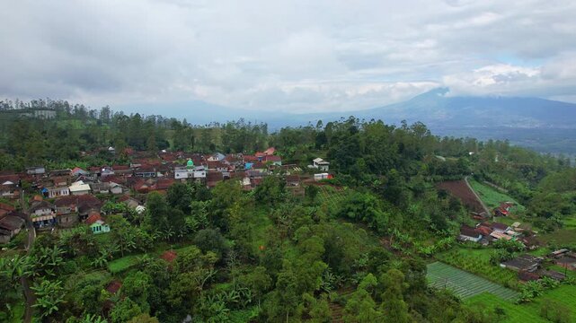 Aerial drone footage of a local village surrounded by forests and trees, on top of a hill, in cloudy atmosphere, with volcano mountains behind, in Garut area, Java island, Indonesia