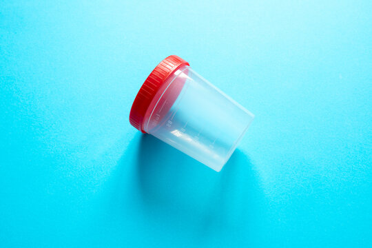 empty, transparent plastic specimen container with red screw-cap lies diagonally at the center of a solid, vibrant blue background. high angle, minimalist aesthetic with plenty of negative space