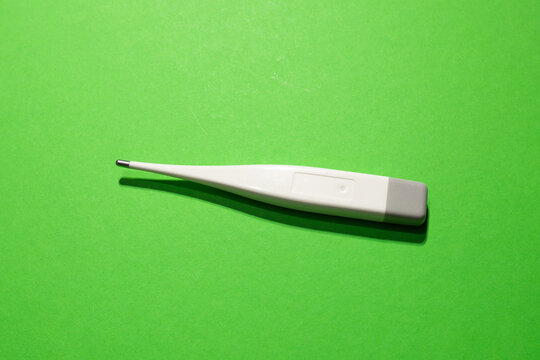 A white digital thermometer lies on a vibrant green background with plenty of empty negative space. A minimalist composition focusing on healthcare and medical equipment