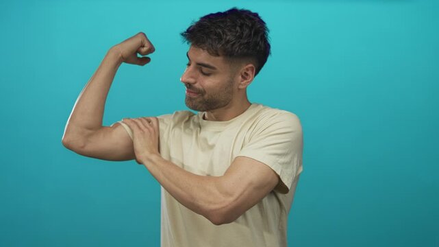 Young hispanic man flexing biceps and smiling with eyes closed in studio against a teal wall; confidence.