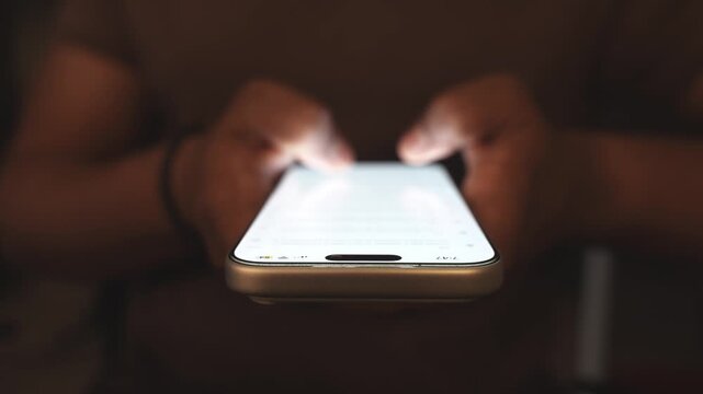 Hand holding smartphone and scrolling social media feed in dark room with desk on background, illuminated by warm tone light. Digital addiction and mindless content consuming.