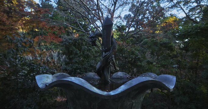 Water coming out of old dragon statue's mouth and into fountain with autumn foliage behind