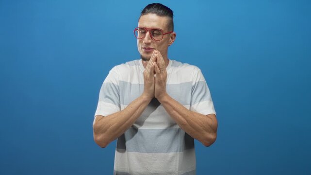 Man with red glasses covers mouth with hands in a blue studio, striped shirt visible and later presses palms together; surprise contemplation.