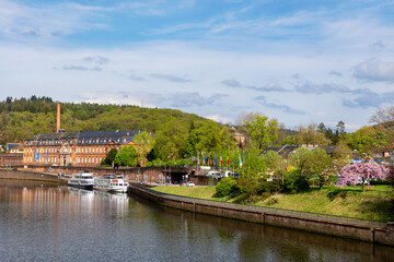 Fototapeta premium Scenic view Mettlach historic architecture industrial heritage Saar river Germany forested hills calm waterfront. Scene panorama cruise boat brick buildings, chimney vibrant spring greenery blue sky