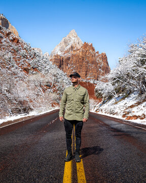 View of a lone figure stands centered on a stark black road bisected by vibrant yellow lines, framed by snow-dusted red rock mountains under a clear blue sky, Springdale, Utah, United States.