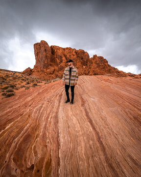 View of a lone figure stands atop a mesmerizing, undulating red rock formation beneath a brooding sky, creating a striking contrast of color and texture, Moapa Valley, Nevada, United States.