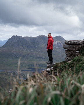 View of a lone figure in a vibrant red jacket stands poised on a rocky precipice, gazing across the rugged, mountainous terrain under a brooding sky, Ullapool, Scotland, United Kingdom.