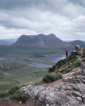 View of a lone figure in a red jacket gazing out over the rippling loch towards the majestic mountain under a brooding sky, Ullapool, Scotland, United Kingdom.