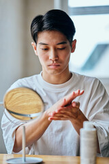 Asian young man applying skincare lotion in front of mirror