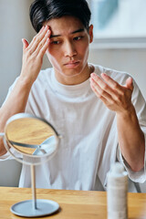 Asian young man applying skincare to forehead in front of mirror