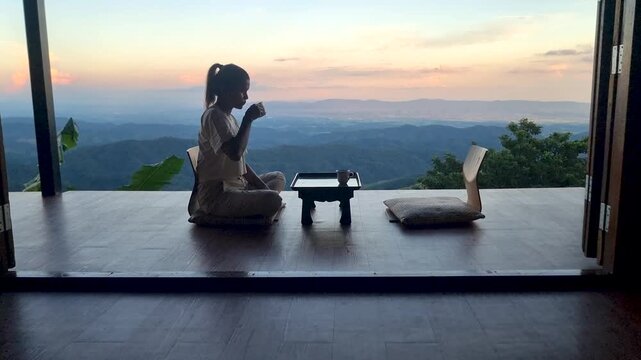 A tranquil moment unfolds as a woman sips tea while sitting on a wooden floor overlooking the breathtaking mountains of Chiang Mai, Thailand at sunset. Natures beauty enhances relaxation and peace.