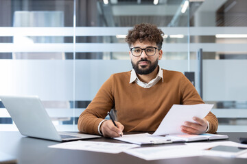 Fototapeta premium Young man with eyeglasses and beard sitting at a desk in a modern office, concentrating on paperwork and using a laptop, representing business analysis and professional work