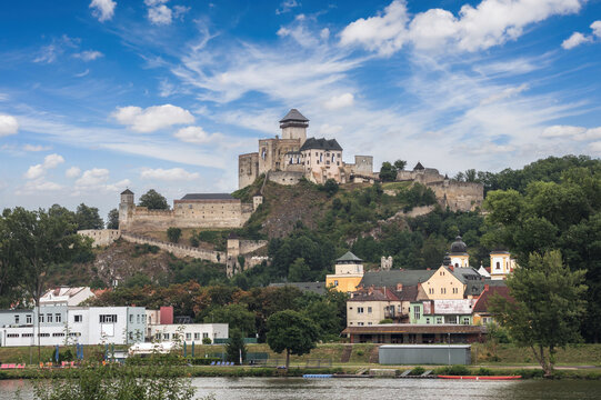 View of medieval Trenc&iacute;n Castle rising above V&aacute;h River and colorful houses on green hillside under blue sky with clouds. Iconic Slovak fortress landscape
