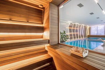 Fototapeta premium Wooden benches are placed inside a sauna room. A view of an indoor pool is seen through large glass windows. A small tray of towels sits on the bench