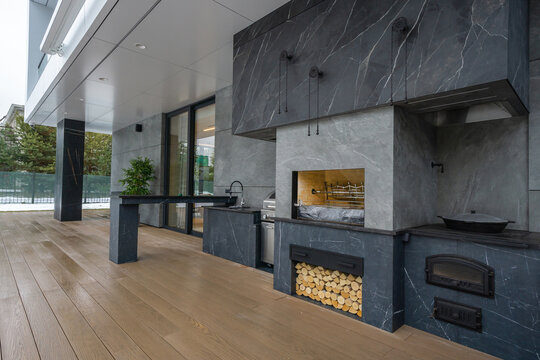 A modern outdoor kitchen is built on a wooden deck. It features a stone cooking area, a prep space, and stacked wood for grilling. Green plants sit nearby, enhancing the area
