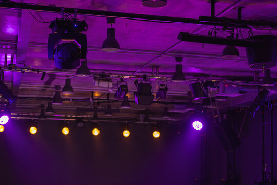 Dramatic purple stage lighting illuminates overhead rigging and spotlights above an empty concert or theater stage, creating moody ambiance for live music, events, production, and entertainment scenes