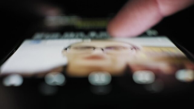 Extreme close-up of a thumb swiping up a touchscreen on a social app news feed in a dark setting. The screen illuminates the finger while scrolling posts photos and video content.