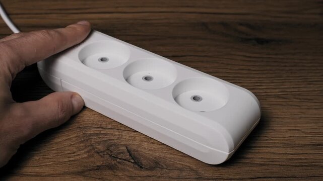 Close-up of a male hand unplugging a black power cord with a European plug from a white triple socket extension block. The setup on a dark wood table disconnects for energy saving.