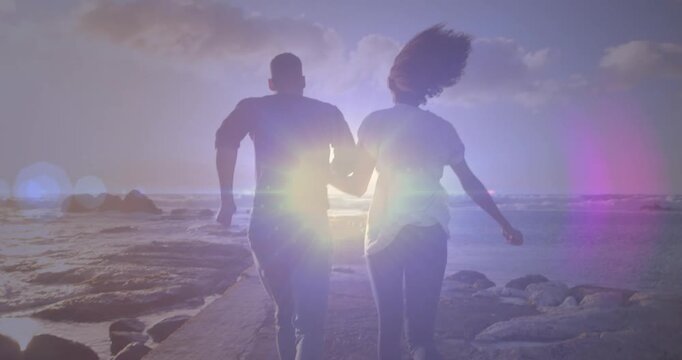 Bright spot of light against rear view of african american couple running on the rocks near the sea